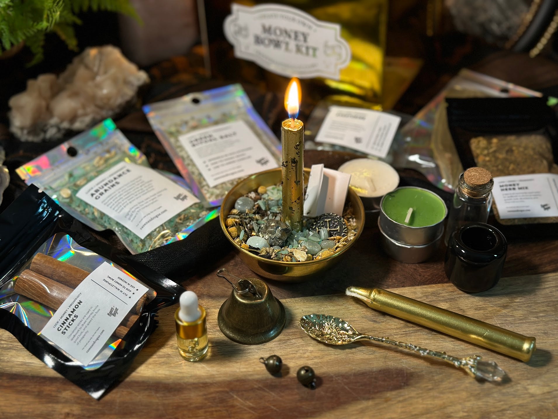 Raven and Rogue Money Bowl Kit product photo. A lit gold taper candle stands in a polished brass bowl filled with aventurine crystals, cinnamon sticks, dried herbs, and gold leaf on a worn wooden surface. A burning tea light and two small brass bells sit nearby. The gold foil Money Bowl Kit box is visible in the background.