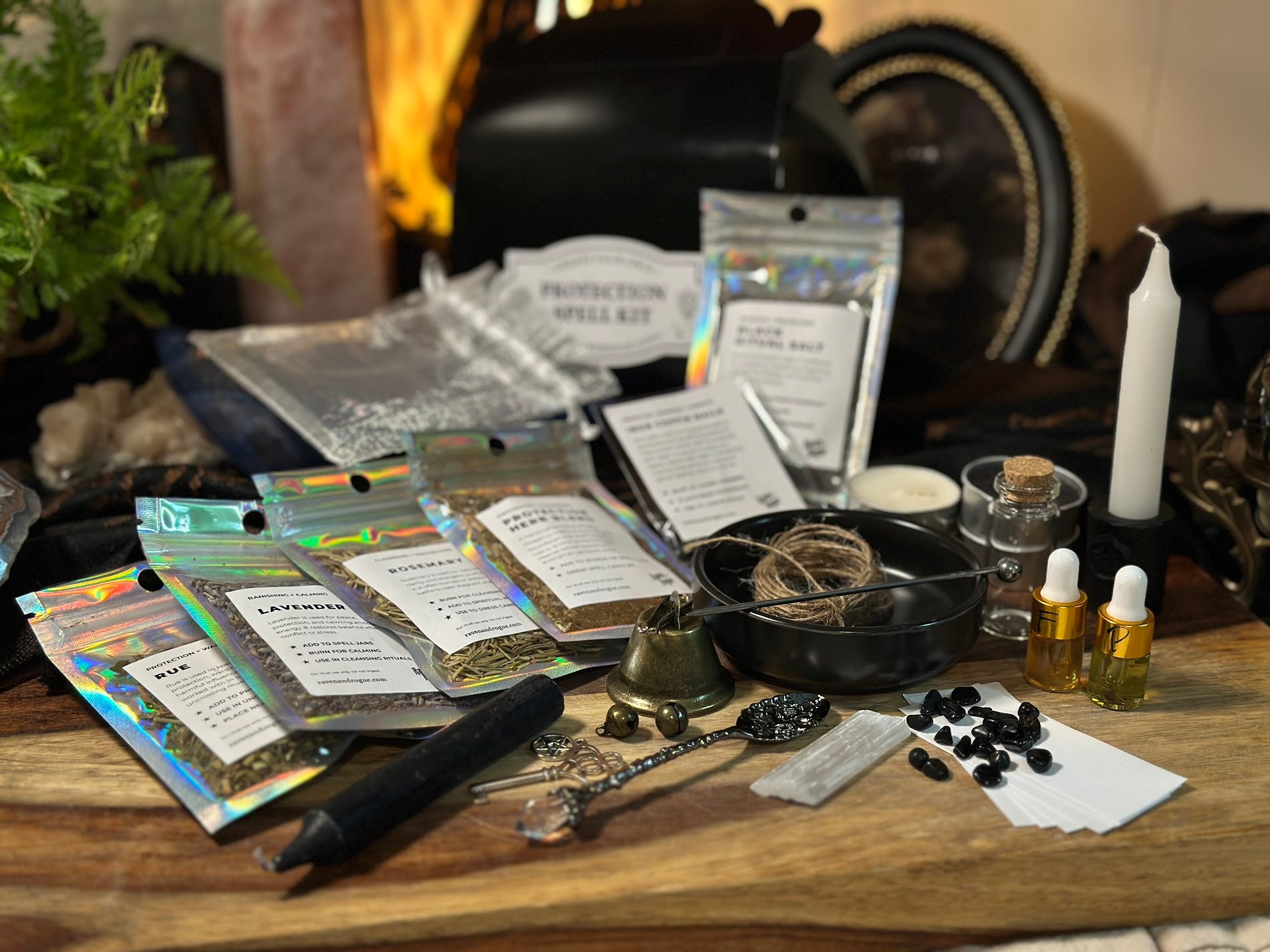 Raven and Rogue Protection Spell Kit components spread on a wooden surface. Holographic pouches labeled Rue, Lavender, Rosemary, and Protection Herb Blend fan out on the left beside a black carving tool. Center shows a black bowl with twine and carving needle, brass bell, key charm, and black tourmaline chunks. Right side holds a white chime candle, tealight, corked bottle, two gold dropper oil vials, selenite wand, and parchment cards.