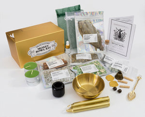 A comprehensive money bowl kit arranged on a clean white background, featuring a gold-colored box, a gold bowl, gold taper candles, green tea lights, herbal blends in clear packaging, a small black jar, wooden tools, and printed instruction/brand cards.