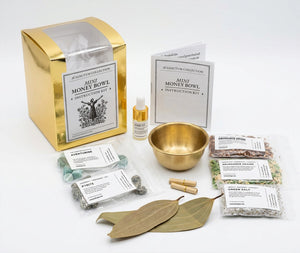 A gold-toned mini money bowl kit arranged on a white background, including a metallic gold box, instruction booklet, small bottle, brass bowl, crystals, salt packets, and two bay leaves.