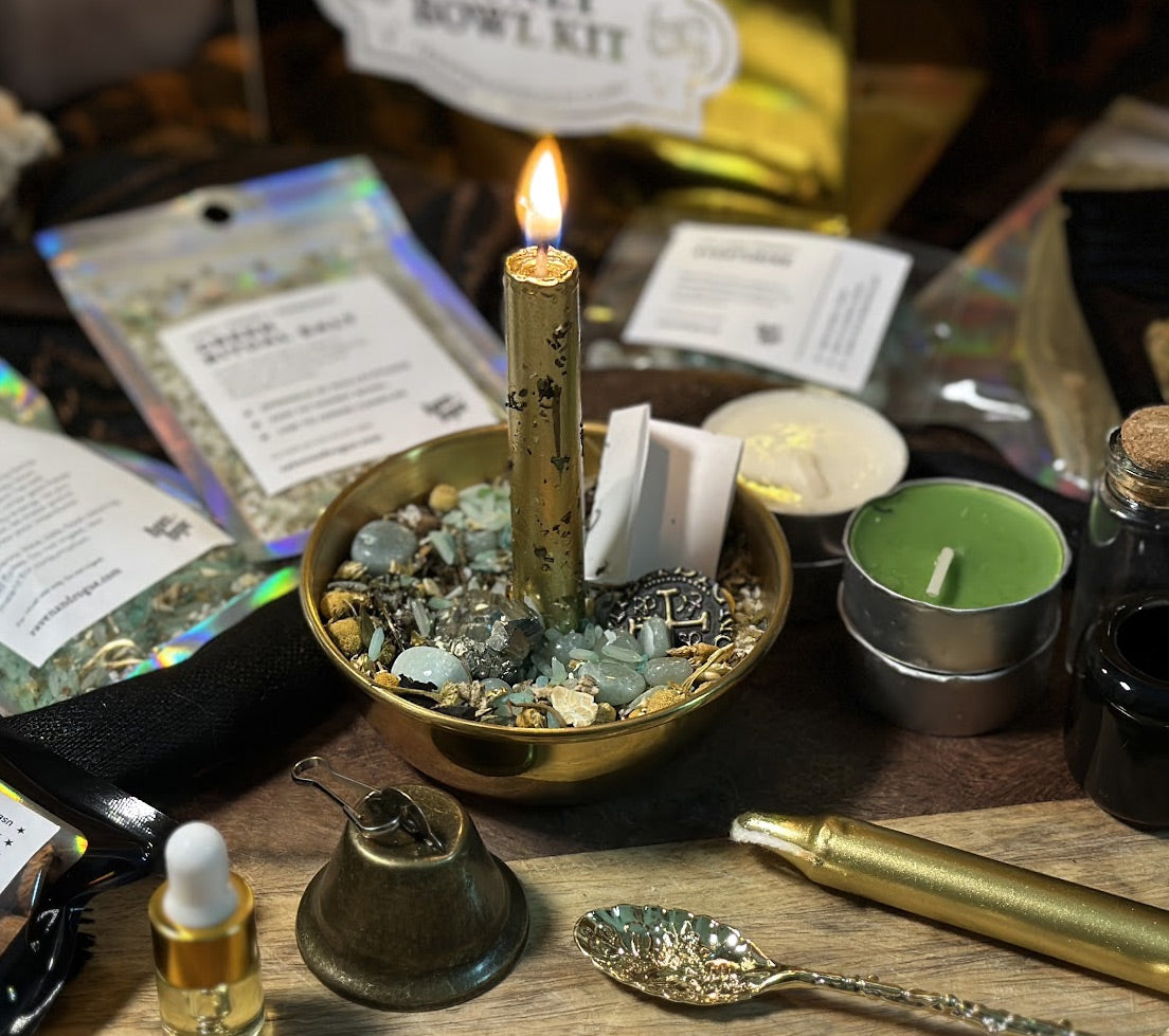 A styled tabletop arrangement for a candle-making or ritual kit, featuring a lit gold taper candle in a brass bowl filled with crystals, surrounded by wax melts, green tea candles, a small brass bell, a gold spoon, and ingredient packets on a wooden surface.