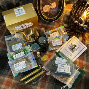 A styled flat-lay of a ritual or candle-making kit arranged on a plaid blanket, including a gold box labeled 'Money Bowl Kit,' several candles, pouches of herbs and wax, a metal bowl, a decorative frame, and a glowing candle holder.