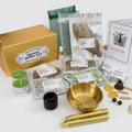 A comprehensive money bowl kit arranged on a clean white background, featuring a gold-colored box, a gold bowl, gold taper candles, green tea lights, herbal blends in clear packaging, a small black jar, wooden tools, and printed instruction/brand cards.