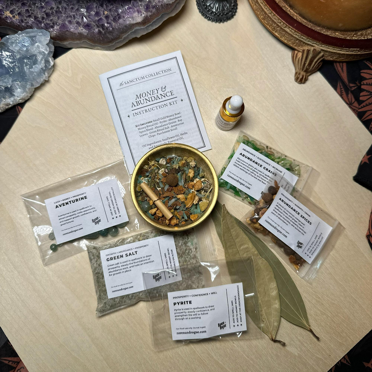 A tabletop flat lay of a miniature money bowl kit by Raven and Rogue. The set includes labeled pouches of herbs and salts, a small dropper bottle of oil, a round gold bowl, crystal chips, bay leaves, and printed cards placed on a light wood surface.