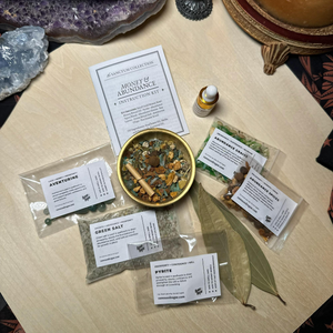 A tabletop flat lay of a miniature money bowl kit by Raven and Rogue. The set includes labeled pouches of herbs and salts, a small dropper bottle of oil, a round gold bowl, crystal chips, bay leaves, and printed cards placed on a light wood surface.
