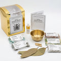 A gold-toned mini money bowl kit arranged on a white background, including a metallic gold box, instruction booklet, small bottle, brass bowl, crystals, salt packets, and two bay leaves.