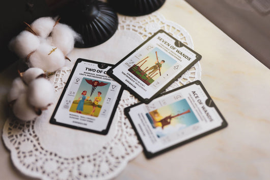 Three-card tarot spread from Tarotorial Expanded deck on white lace cloth. Hand with dark painted nails holds cotton plant while cards show Two of Cups (partnership/love), Seven of Wands (perseverance/challenges), and Ace of Pentacles (new opportunities/manifestation). Soft natural lighting creates intimate reading atmosphere.
