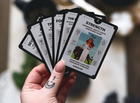 Hand holding fanned Tarotorial Expanded tarot cards showing multiple cards with Strength visible. Cards feature clean design with artwork, keywords, and astrological symbols. Blurred indoor background with soft lighting.