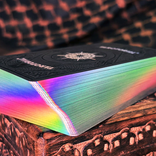 Stack of Tarotorial Expanded tarot cards showcasing vibrant holographic rainbow edges. The black card backs feature ornate compass design with "TAROTORIAL" text. Cards rest on textured wooden surface with warm ambient lighting.