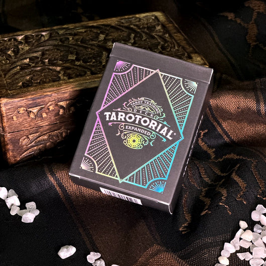 Tarotorial Expanded mini deck box on dark wooden surface. The black box features vibrant holographic foil design with geometric patterns in pink, teal, and gold. The Tarotorial logo and "Expanded" text are prominently displayed with decorative compass-style artwork.