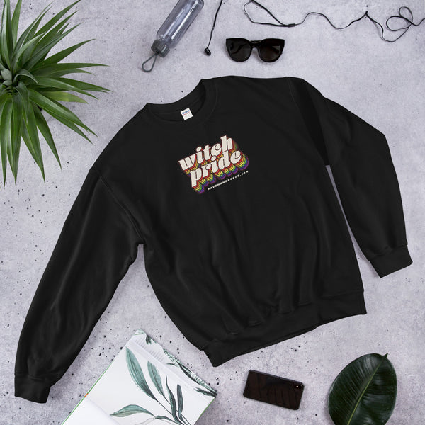 A black sweatshirt with a design that reads "Witch Pride" on it sits on a concrete floor. Surrounding the sweatshirt are sunglasses, a water bottle, a plant, a phone, and a journal.
